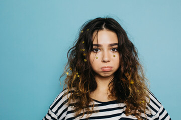 Sad sore resentful young woman with puffy cheeks cover with golden star-shaped glitter. Attractive woman with curly brown hair looking at camera. Isolated over blue background.