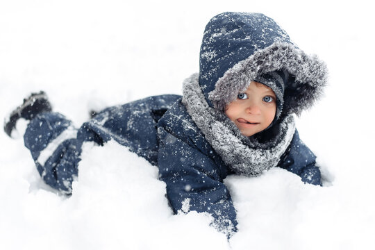 Close-up image of cute baby boy in a warm white snow suit lying in snow with funny face.
