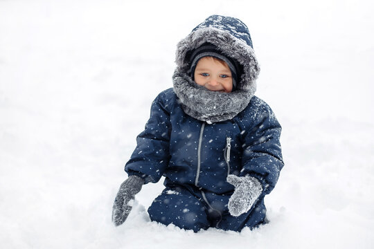 Image of positive baby boy in a warm white snow suit sitting on snow.