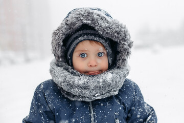 Surprised baby boy in a warm white snow suit cathing snow with tongue.