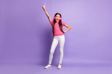 Full length body size photo of small girl smiling listening to music pointing with finger laughing isolated on purple color background