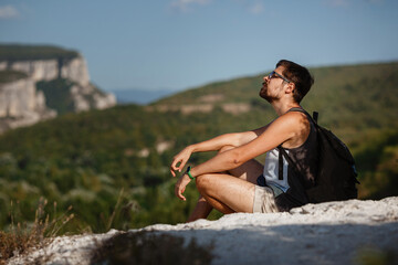 Young male hiker with backpack relaxing on top of a mountain during calm summer sunset