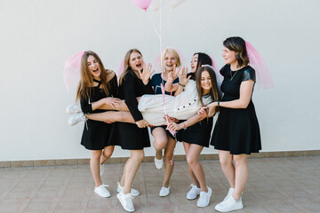 Fototapeta premium Bride and bridesmaids stands near white wall outdoors and celebrating bachelorette party. Beautiful sexy young girls in dress and veil on bachelor party. Maiden evening, Hen-parties. Place for text.