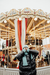Obraz na płótnie Canvas Beautiful girl in stylish fashion jacket and knitted hat and scarf happy covers his face with his hands near the carousel in amusement park.