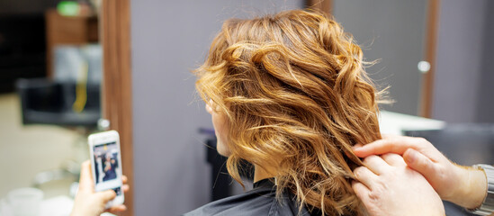 Fototapeta premium Hairdresser doing hairstyle for young woman with red curly hair and with smartphone in her hands in beauty salon