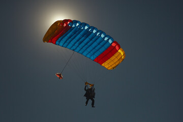 The bright multi-colored parachute canopy with the silhouettes of the instructor and passenger tandem against the dark blue sky illuminated by the dazzling sun, close-up.