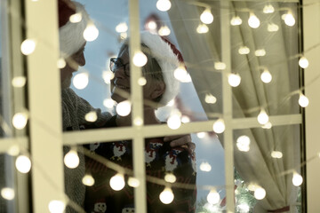 Elderly couple with Santa hats stand indoors behind the lighted window - Merry Christmas and expression of love for two retired seniors enjoying the nativity holidays