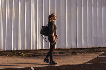 Fitness woman in black tracksuit outdoor, white wall background. Woman with gym bag, wireless headphones and water bottle in hand. Sunset light, full length.