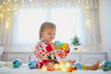 Happy little toddler girl wearing pajamas and playing with Christmas decorations on bed