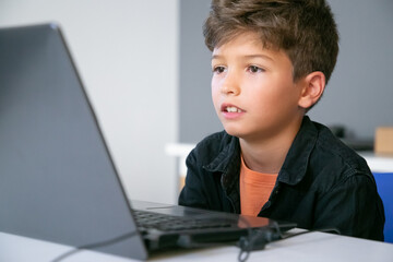 Caucasian boy sitting at table in classroom, reading text on screen or watching video presentation. Concentrated pupil studying in computer school. Knowledge and digital education concept