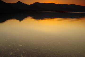 夕暮れの湖。湖面に映る黄昏の空と山並みのシルエットから成る静寂の印象。