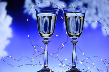 Two glasses of champagne against the background of a winter fairy-tale landscape. Glittering lights and reflection in a glass of fir trees in the snow. Christmas background concept.