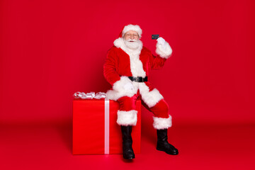 Full length body size view of his he handsome fat overweight Santa sitting on large big giftbox holding in hand plastic card isolated bright vivid shine vibrant red color background