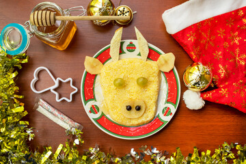 Festive Bull-shaped porridge for 2021 on wood background, christmas tree toy, honey, sprinkles, bakeware