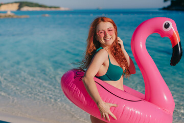 A young happy female in a swimsuit is standing and holding an inflatable pink flamingo on the beach...