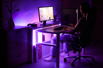 Gamer sitting at the table in front of computer monitor and playing computer game in dark room at home