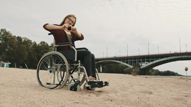 Suport for disabled people. Young woman in the wheelchair on the sandy river coast showing heart with hands. High quality 4k footage