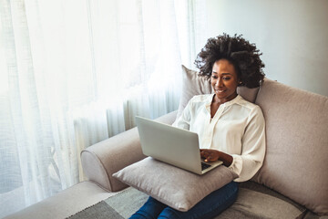 Young woman video calling using a laptop sitting on a sofa wearing earphones. Happy african american young business woman wearing headphones, waving hello.