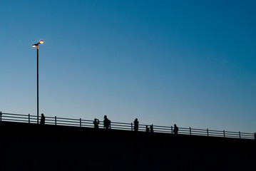 Silhouette of people walking across Waterloo Bridge, photographed at dusk with a single lit streetlight in shot. South Bank, London, UK. Horizontal.