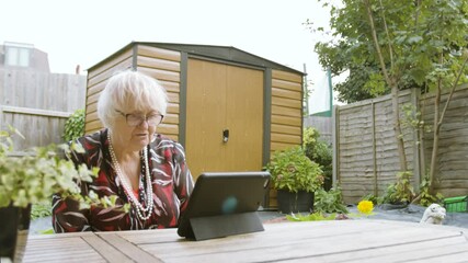 Elderly Woman speaking on zoom on a tablet device laughs and shrugs in garden