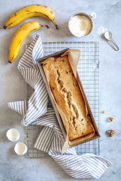 Flat lay with banana bread on a cooling rack with ingredients laying around including eggs, bananas, walnuts and flour on white background