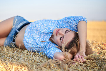 Young curly blond woman, wearing jeans shorts and light blue shirt, lying on bale on field in...