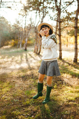  Stylish woman with a beautiful smile at sunset in the forest. Hipster girl is dressed with a hat, a light sweater and a skirt. The concept of youth, love, autumn and lifestyle.
