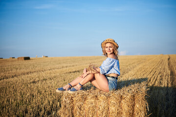 Naklejka na ściany i meble Young curly blond woman, wearing jeans shorts, light blue shirt and straw hat, sitting on bale on field in summer. Female portrait in natural rural scene. Environmental eco tourism concept.