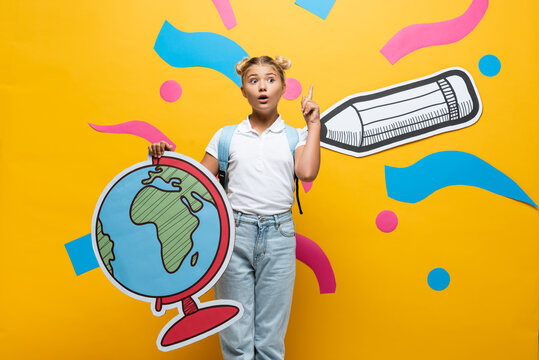 amazed schoolgirl showing idea gesture while holding globe maquette near paper pencil and colorful elements on yellow