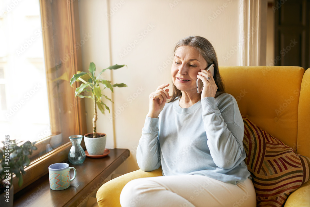 Poster beautiful female pensioner wearing casual clothes relaxing at home, sitting in armchair by window, d