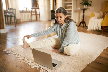 Indoor image of flexible active mature gray haired female sitting on floor in front of open laptop, stretching legs during online yoga lesson, watching live video with professional instructor