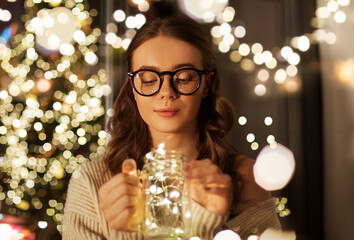 christmas, holiday and people concept - young woman in glasses with festive garland lights in mason jar mug at home