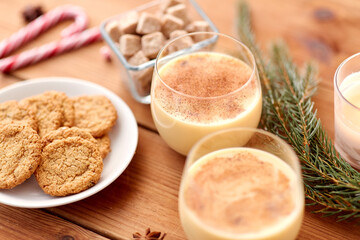 christmas and seasonal drinks concept - glasses of eggnog with oatmeal cookies, fir tree branches, candy canes and sugar on wooden background