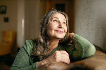 Close up image of beautiful positive pensive European female pensioner with gray hair and wrinkled skin relaxing at home on comfortable leather couch, looking up with dreamy thoughtful smile