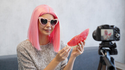 Positive blogger records a video about healthy eating and holds a watermelon in her hands. Woman wearing pink wig and heart shaped glasses