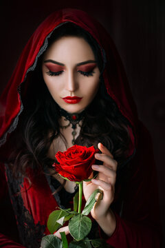 Mystical beautiful woman in a gothic costume of a medieval vampire in a hood. An adult girl holds a rose flower in her hands. Beautiful face red makeup, attractive lips. Halloween party image.
