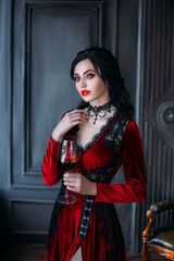 Beautiful young sexy woman vampire in medieval dark castle. Red gothic evening dress. Black wavy hair. Backdrop vintage room. A girl holding a glass of bloody wine in her hands. Image queen of night  © kharchenkoirina