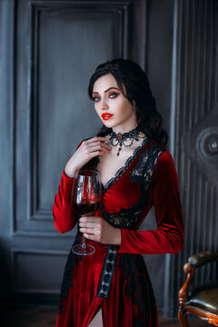 Beautiful young sexy woman vampire in medieval dark castle. Red gothic evening dress. Black wavy hair. Backdrop vintage room. A girl holding a glass of bloody wine in her hands. Image queen of night 