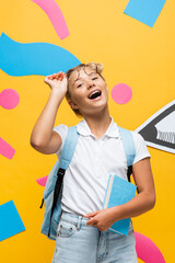 excited schoolgirl touching eyeglasses while holding book on yellow background with paper pencil and decorative elements