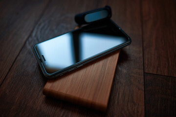 A cell phone and a smart bracelet lie on a wooden table stand. The light from the window hits the screen and the bracelet.