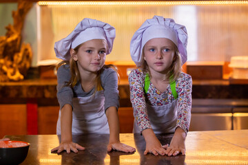 Little girls in white chef uniform and hats cooking by the table in kitchen. Two cute sisters preparing dough