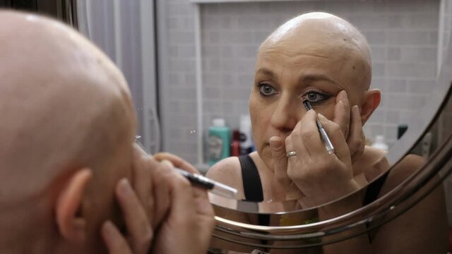 bald woman with cancer puts on makeup in front of the mirror