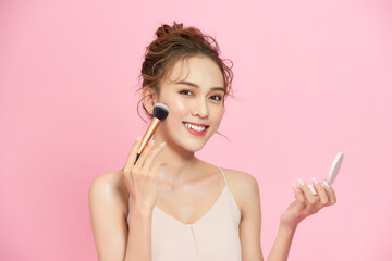 Closeup portrait of beautiful happy young girl with perfect skin is holding makeup brush in hand. Isolated on pink