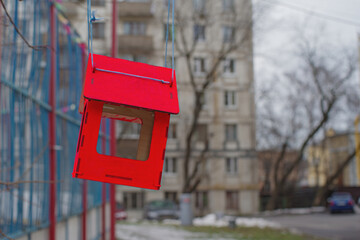 Bright red bird feeder hanging on a tree branch in a city courtyard. Children's сare for birds in difficult winter times.