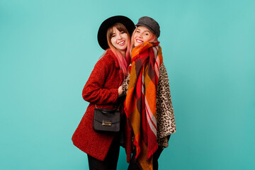 Two stylish blond  women posing in studio on blue turquoise background. Friends hugging and having fun together.  Wearing leopard print faux fur coat and wool scarf. Winter fashion.