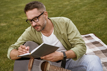 Writing notes. Young focused man wearing eyeglasses holding acoustic guitar and composing a song while sitting on a green grass outdoors, male musician working in park