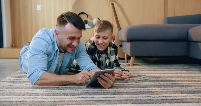 Close up of good-looking happy smiling father and his son which lying on the floor in the living-room and using i-pad to watch interesting apps