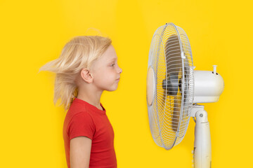 Blond boy stands near ventilator on bright yellow background. Hot summer.