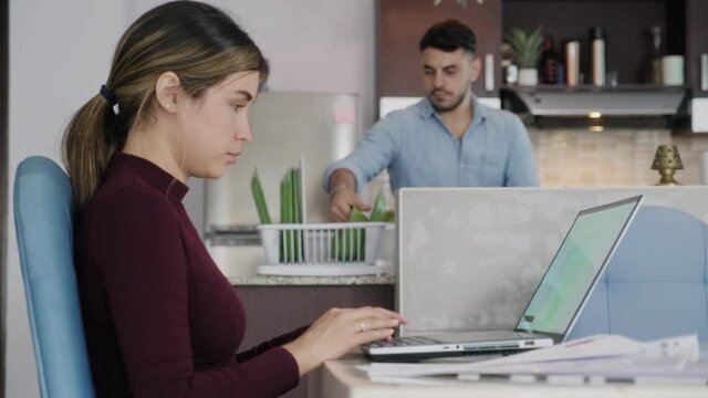 Multiethnic couple at home: busy wife is working with laptop computer, husband is doing chores. Hispanic business woman typing on office pc and sending email for remote work, white man washing dishes
