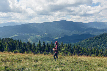 Fototapeta na wymiar Girl with pink hair climbs uphill mountain. Hiking through forest in summer. Standing on the clearing and looks at the mountains. Dark autumn forest. Local Travel
